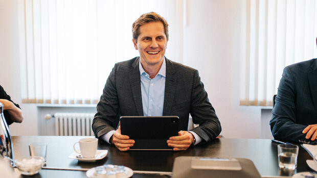 dieses Bild zeigt eine Personen, die an einem Tisch sitzt und die gegenüber sitzenden Personen anlächelt