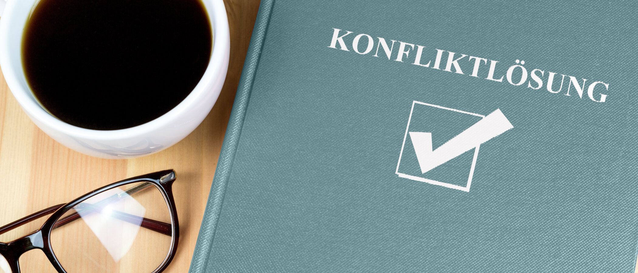 dieses Bild zeigt eine Tasse mit Kaffee, eine Brille und ein Buch, auf dem das Wort Konfliktlösung steht