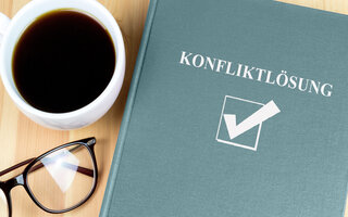 dieses Bild zeigt eine Tasse mit Kaffee, eine Brille und ein Buch, auf dem das Wort Konfliktlösung steht